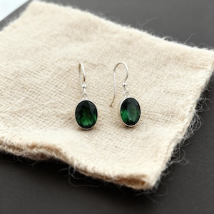 Puede incluir: Un par de pendientes colgantes de plata con piedras preciosas ovaladas de color verde oscuro. Los pendientes se muestran sobre una tela texturizada de color blanquecino, sobre un fondo oscuro. Las piedras tienen una superficie pulida y reflectante.