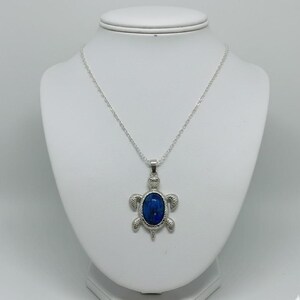 Può includere: Una collana color argento con un ciondolo a forma di tartaruga. Il ciondolo presenta una grande pietra ovale blu scuro incastonata in una cornice a forma di tartaruga. La collana è esposta su un busto bianco.
