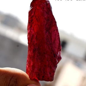 May include: A deep red, rough-cut gemstone with a translucent appearance. The gemstone is held in a hand.