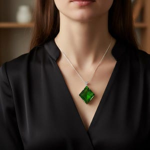 Puede incluir: Un collar de plata con un gran colgante de piedra preciosa verde esmeralda de corte cuadrado. El colgante cuelga de una delicada cadena de plata. La mujer lleva una blusa negra.