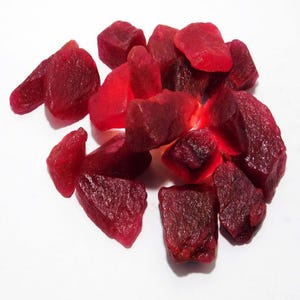 Könnte beinhalten: Ein Haufen roher, roter Rubin-Edelsteine. Die Steine sind in verschiedenen Größen und Formen, einige haben einen polierteren Look.