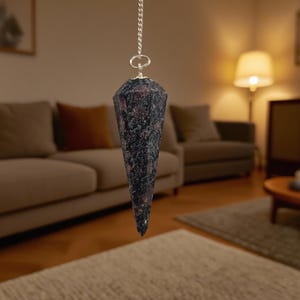 May include: A faceted, cone-shaped pendulum with a silver chain. The pendulum is made of a dark stone with a speckled pattern of black, gray, and red. It is suspended in front of a blurred living room background.