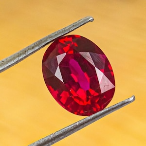 Puede incluir: Una gema ovalada de color rojo vibrante sostenida por unas pinzas. La gema tiene un corte facetado y un color rojo intenso.