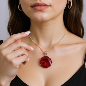 Puede incluir: Un collar dorado con un gran colgante redondo rojo. El colgante cuelga de una delicada cadena. También se ven pendientes de aro dorados. La joya se lleva sobre un top negro.