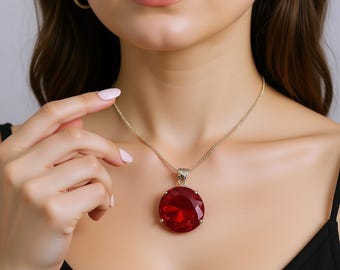 Op de afbeelding: Een goudkleurige ketting met een grote, ronde, rode hanger. De hanger hangt aan een fijne ketting. Bijpassende gouden oorringen zijn ook zichtbaar. De sieraden worden gedragen op een zwarte top.