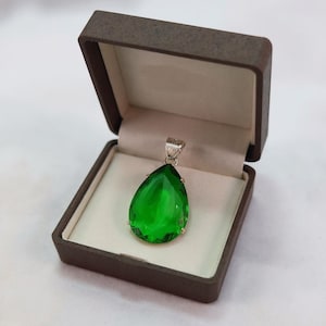 Pode incluir: Um pingente verde vibrante em forma de gota com uma argola prateada, exibido em uma caixa de joias marrom aberta. As facetas do pingente capturam a luz, mostrando sua clareza e cor. A caixa tem um interior creme.