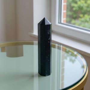 May include: A tall, black crystal obelisk with a faceted design. The polished stone stands upright on a round table with a gold-colored metal frame and a glass top. The background includes a window and natural light.