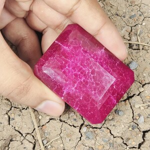 May include: A large, rectangular, faceted gemstone in a vibrant magenta hue. The stone has a complex internal pattern and is held in a hand, showcasing its size and colour. The gem is set against a backdrop of cracked earth.