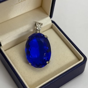 Peut inclure: Un grand pendentif ovale bleu profond avec une bélière et des griffes argentées, présenté dans un écrin bleu à l'intérieur crème. Les facettes du pendentif captent la lumière, créant un effet scintillant.