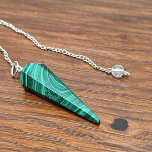 Può includere: Un pendolo in malachite verde sfaccettata, con una catena d'argento e una perla di cristallo trasparente. Il pendolo è a forma di cono e presenta un motivo naturale a strisce. La catena è attaccata a un piccolo anello d'argento.