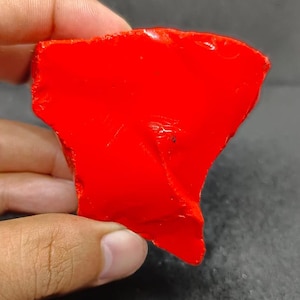 Puede incluir: Una pieza de vidrio rojo brillante de forma irregular. El vidrio tiene una superficie lisa y pulida.