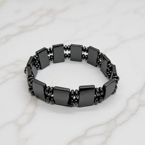May include: A dark gray bracelet featuring rectangular and round beads. The bracelet is made of a shiny, metallic material and is designed to be worn around the wrist. The beads are evenly spaced and connected.