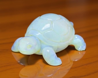 May include: A small, opalescent turtle figurine with a light blue and white color scheme. The turtle has a detailed shell pattern and is resting on a reflective surface. The figurine is a decorative item.