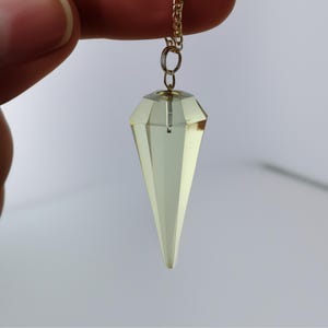 Puede incluir: Un péndulo de cristal amarillo pálido facetado, suspendido de una delicada cadena dorada. El cristal tiene forma de diamante en la parte superior, que se estrecha hasta un punto. La cadena está sujeta por los dedos de una persona sobre un fondo liso.