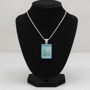 May include: A rectangular pendant necklace with a silver chain. The pendant has a shimmering, iridescent surface with shades of blue, green, and purple. The necklace is displayed on a black jewelry bust.