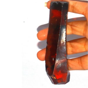 Può includere: Una grande gemma sfaccettata di colore rosso scuro con una punta appuntita. La gemma è tenuta in una mano.