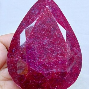 May include: A large, deep red ruby gemstone with a pear-shaped cut. The gemstone is faceted and has a high degree of brilliance.