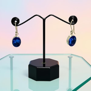 May include: A pair of silver-toned earrings with large, oval, deep blue gemstones. The earrings are displayed on a black jewelry stand with a hexagonal base. The gemstones are the focal point, catching the light.
