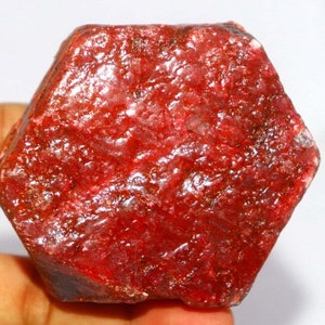 May include: A raw, uncut ruby gemstone in a deep red hue. The ruby has a hexagonal shape and a textured surface, displaying its natural, unpolished form. The gemstone is held in a hand against a white background.