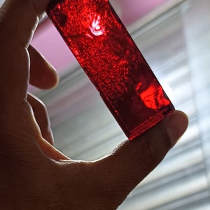 Puede incluir: Un bloque rectangular de resina transparente de color rojo oscuro con burbujas de aire atrapadas en su interior. La resina es translúcida y tiene un acabado brillante.