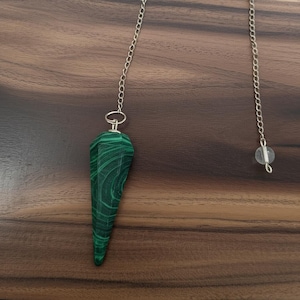 May include: A green malachite pendulum with a silver chain and a clear crystal bead. The pendulum has a pointed, faceted shape and a distinctive banded pattern. The chain is attached to the top of the pendulum.