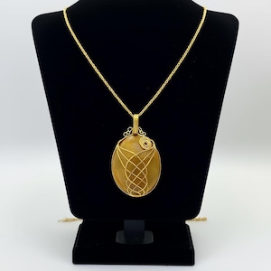 Op de afbeelding: Een goudkleurige hangerketting met een ovaalvormige, amberkleurige steen, omwikkeld met een geweven gouddraadontwerp. De hanger hangt aan een delicate gouden ketting. De ketting wordt gepresenteerd op een zwarte sieradenstandaard.