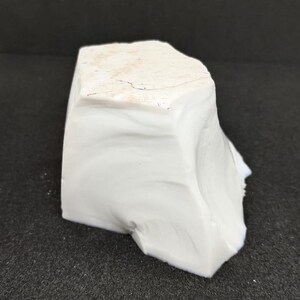May include: A solid, irregularly shaped, white rock with a slightly textured surface. The rock has a light beige top and a smooth, opaque appearance. The object is set against a dark background.
