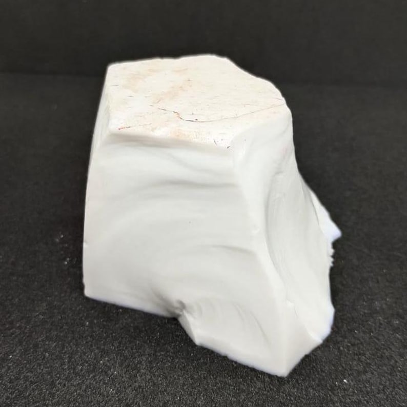 May include: A solid, irregularly shaped, white rock with a slightly textured surface. The rock has a light beige top and a smooth, opaque appearance. The object is set against a dark background.