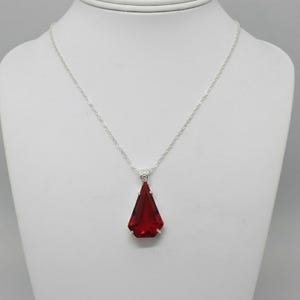 May include: A silver necklace with a pendant featuring a large, faceted, deep red gemstone. The pendant is a triangular shape with a silver setting. The necklace is displayed on a white mannequin.