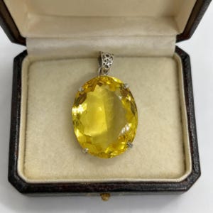 Può includere: Un grande pendente ovale in pietra preziosa gialla con una cauzione color argento. Il pendente è incastonato in un metallo color argento ed è esposto in un portagioie marrone con interno color crema. La gemma ha molte sfaccettature.