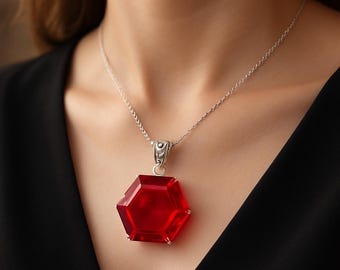 Op de afbeelding: Een zilveren ketting met een grote, rode, zeshoekige hanger. De hanger is facetgeslepen en hangt aan een fijne zilveren ketting. De ketting wordt getoond tegen een donkere achtergrond, waardoor de rode edelsteen wordt benadrukt.