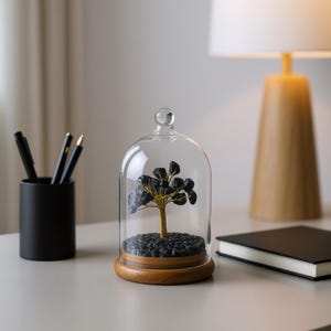 May include: A decorative glass cloche with a wooden base houses a miniature tree with black stone leaves and a gold-colored trunk. The cloche sits on a light gray surface, next to a black pen holder and a wooden lamp.
