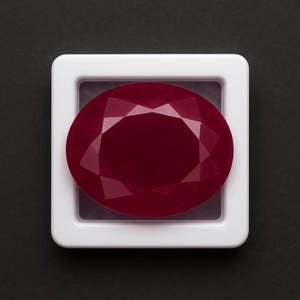 May include: A faceted, oval-shaped ruby gemstone, displayed in a white plastic case against a black background. The ruby is a deep, rich red color, with multiple facets that catch the light.