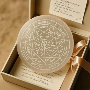 May include: A round, off-white selenite crystal plate with etched geometric patterns and names like Raphael and Michael. The plate is in an open box with a cream-colored ribbon and handwritten notes.