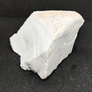 May include: A large, irregularly shaped piece of white stone with a slightly textured surface. The stone has a few small, colored inclusions and a light tan area on the top. The stone is set against a dark background.