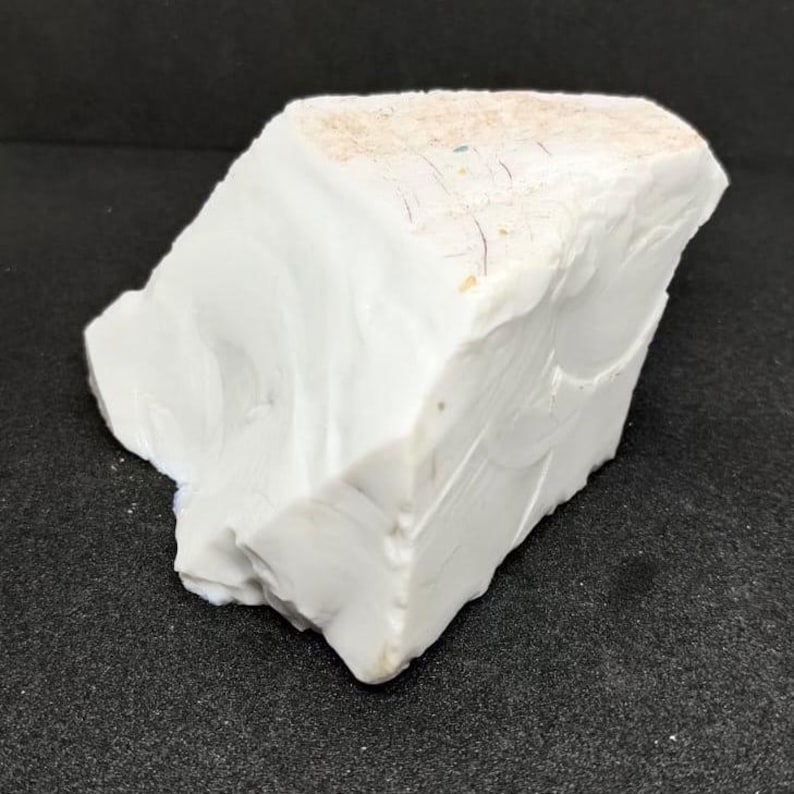 May include: A large, irregularly shaped piece of white stone with a slightly textured surface. The stone has a few small, colored inclusions and a light tan area on the top. The stone is set against a dark background.