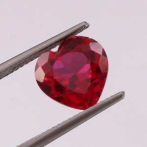 May include: A heart-shaped ruby gemstone with a deep red color. The gemstone is held in tweezers.