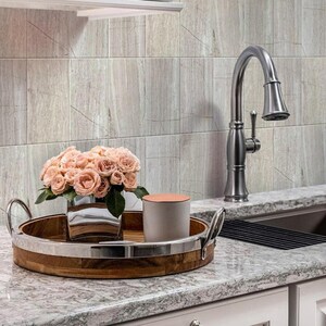 May include: A kitchen scene featuring a stainless steel faucet and a wooden tray with a silver rim. The tray holds a vase of pink roses and a gray candle. The background has light gray tiles.