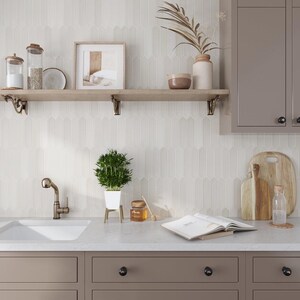 May include: A white kitchen with a white subway tile backsplash, a wooden shelf with a plant, a picture frame, and a glass jar. The kitchen also has a stainless steel sink and a brown countertop.