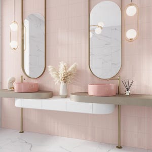 May include: Two oval mirrors with gold frames hang above a double vanity with two pink ceramic sinks. The vanity has a white countertop and a gold metal base. The walls are tiled in a light pink color with vertical grooves.