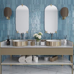May include: A bathroom vanity with a gold metal frame and a white marble countertop. Two oval mirrors with gold frames hang above the vanity. Two gold metal sinks are on the countertop. A bouquet of white flowers is in a gray metal container on the countertop. Two gold metal soap dispensers are on the countertop. Two gold metal candle holders are on the countertop. Two white towels are on the bottom shelf of the vanity.