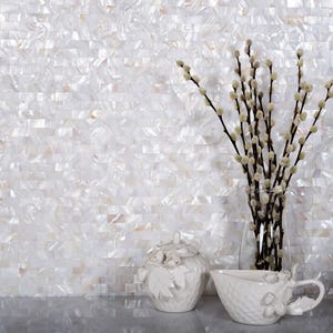 May include: White mother of pearl tile mosaic with a white ceramic sugar bowl and creamer set with floral accents. The background is a white wall with a vase of white branches.