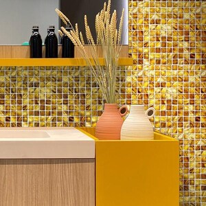 May include: Two ceramic vases, one peach and one white, sit on a yellow shelf in front of a wall covered in small, gold mosaic tiles. A bouquet of wheat stalks sits in the peach vase.