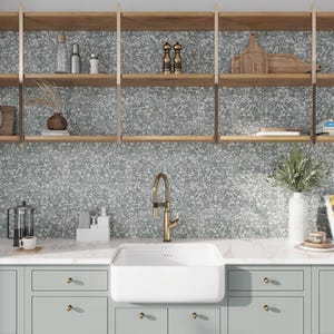 May include: A white farmhouse style kitchen sink with a brushed gold faucet. The sink is installed in a countertop with light green cabinets below. The backsplash is a light gray and white speckled tile.