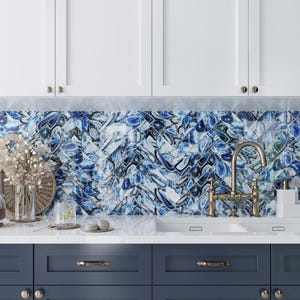 May include: A kitchen with white cabinets and blue patterned tile backsplash. A double sink with a brass faucet is centered in the image. The lower cabinets are dark blue with gold handles.