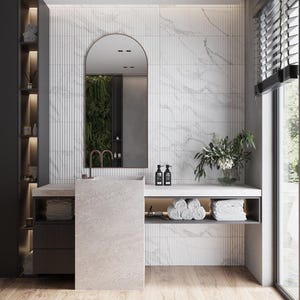 May include: A modern bathroom vanity with a white marble tiled wall, a large arched mirror, and a white stone countertop. The vanity has a single sink and a shelf below for storage. There are two black bottles of soap on the counter and a vase of white flowers on the shelf.