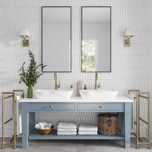 May include: A bathroom vanity with two sinks, two mirrors, and a shelf for towels and toiletries. The vanity is made of light blue wood and has gold hardware. The walls are tiled with white subway tiles.