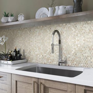 May include: A modern kitchen featuring a stainless steel sink, a chrome faucet, and light-colored wooden cabinets. The backsplash is made of small, round, beige tiles. A shelf above holds white dishes and decorative items.