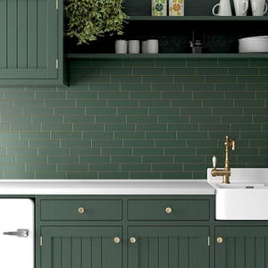 May include: A kitchen with green cabinets and a white countertop. The backsplash is made of green subway tiles with gold grout. There is a white porcelain sink with a gold faucet.