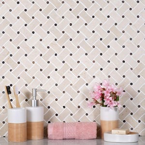 May include: A wall covered in a beige and white basketweave marble tile pattern with small dark brown circular accents. In front are two white and tan toothbrush holders, a soap dispenser, a pink towel, a vase with pink flowers, and soap.